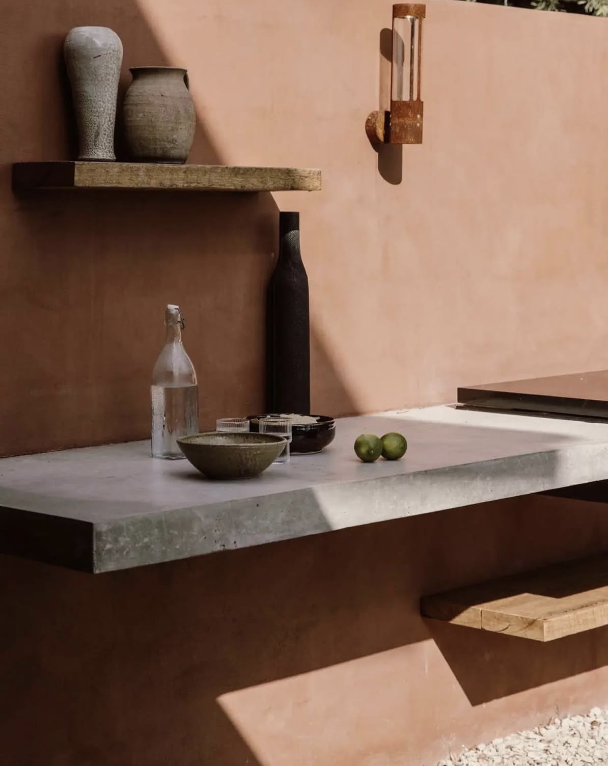 Terracotta outdoor kitchen wall with a floating concrete bench, ceramic vessels on a timber shelf and strong afternoon shadow