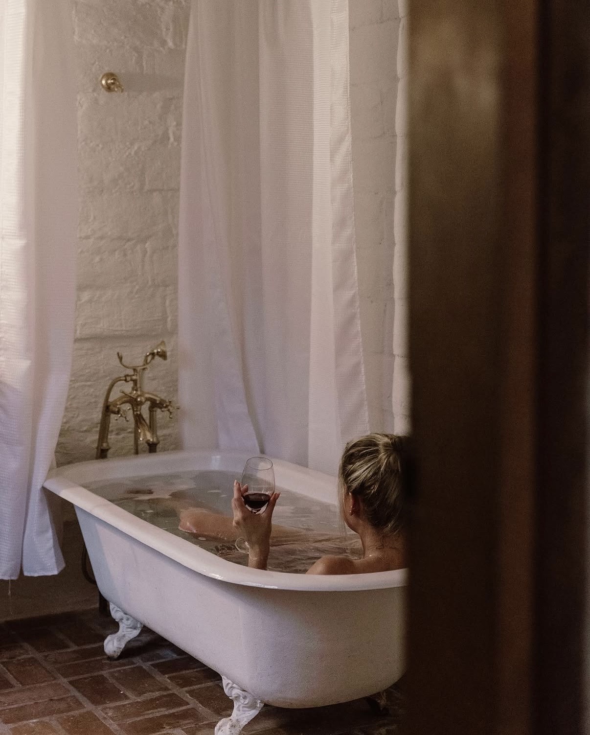 Woman in a freestanding clawfoot bath with brass tapware, sheer curtains and brick flooring